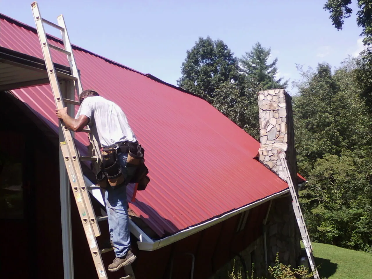 Roofer on a ladder beside a red metal roof and stone chimney — Soffit & Fascia Repair by Roofing Brothers in Paoli
